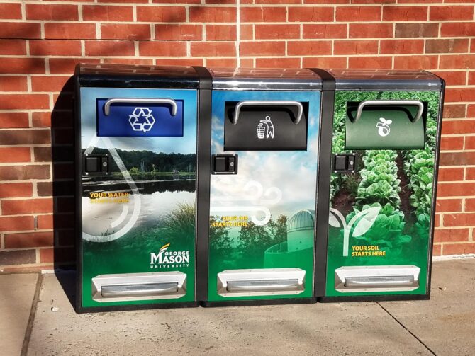 Three large bins with pedal opening, standing side by side against a brick wall. They display symbols indicating that they are for (from left to right) recycling, trash, and composting.