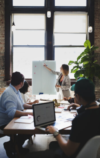 Businesswoman writing on a board in a meeting room while several people seated around a table are watching
