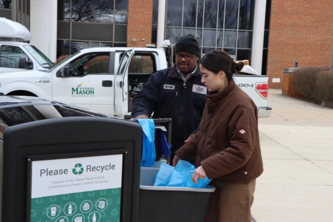 A large recycling bin in the foreground. The inner containers are pulled out and a man and a woman are arranging liners inside. A Facilities van and a brick building with large windows are in the background.